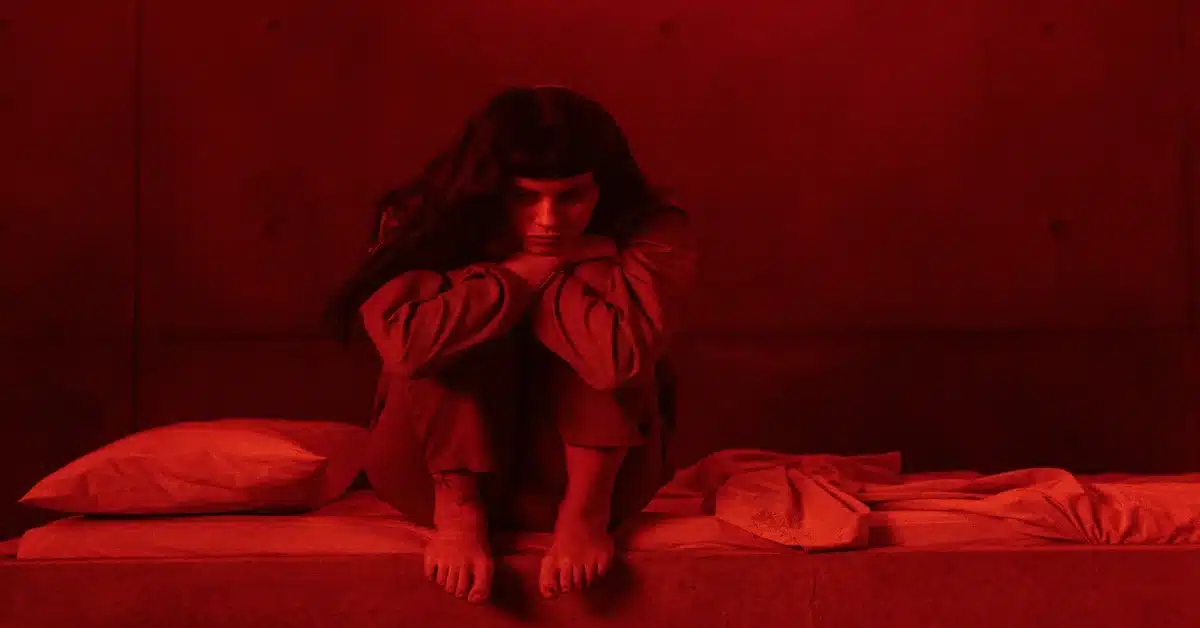 Descrição da imagem: personagem sentada na cama. Imagem: Divulgação.