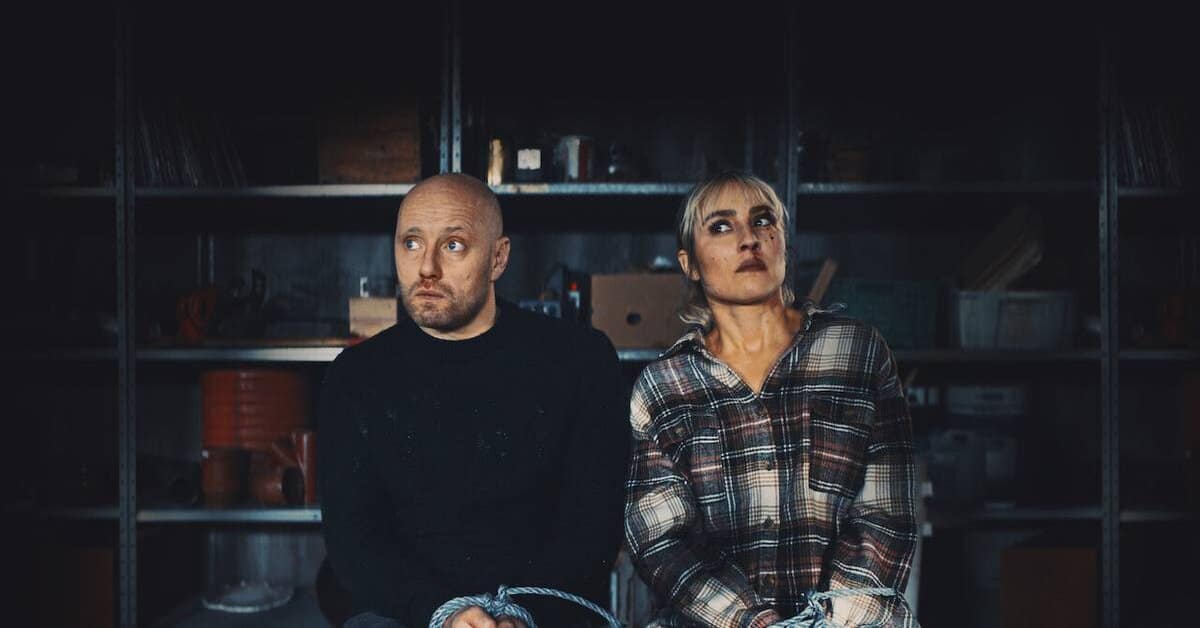 Descrição da imagem: da esquerda para direita, um homem careca e uma mulher amarrados nas mãos em um depósito. Imagem: Divulgação/ Netflix.